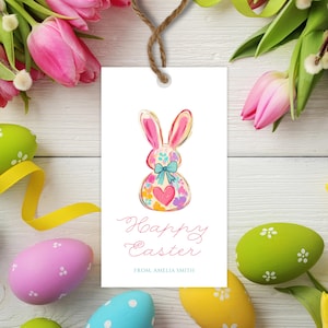 May include: A white gift tag with a pink Easter bunny illustration. The bunny is decorated with colorful flowers and a blue bow. The text "Happy Easter" is written in pink cursive script. The tag also includes the text "From, Amelia Smith".