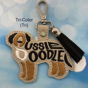 May include: A beige leather keychain with a black and brown embroidered Australian doodle dog design. The keychain has a silver clasp and a black tassel.