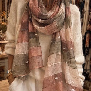 Könnte beinhalten: Ein leichter, rechteckiger Schal mit einem Patchwork-Design aus gedeckten rosa, altrosa und olivgrünen Feldern. Der Stoff hat ein halbtransparentes, strukturiertes Gewebe mit horizontalen goldenen Nähten und kleinen, erhabenen weißen Stickereien.