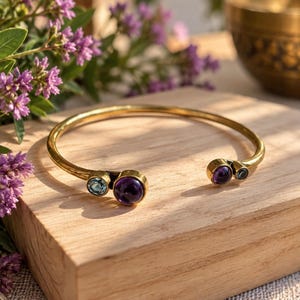 Può includere: Un bracciale rigido aperto color oro con due incastonature circolari su ciascuna estremità. Un lato presenta una grande ametista rotonda viola accanto a un topazio blu, mentre l'altro lato mostra un'ametista viola più piccola e una piccola pietra blu.