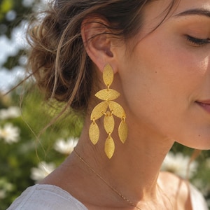 Può includere: Orecchini pendenti color oro con finitura strutturata. Gli orecchini presentano un design a strati con elementi a forma di foglia, sospesi da un punto centrale. È visibile anche una delicata collana d'oro.