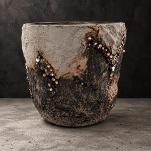 May include: A cylindrical planter with a rough, textured surface featuring a two-tone color scheme of light gray on the upper portion and dark charcoal on the lower section, accented by a diagonal line of embedded black and bronze crystals.