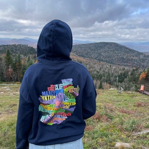 May include: A navy blue hoodie with a colorful graphic on the back. The graphic is a map of a region with the names of different locations printed on it. The person wearing the hoodie is standing in front of a mountain range.
