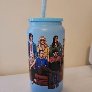 May include: A light blue tumbler with a lid and straw featuring a colourful graphic illustration of the cast of The Big Bang Theory. The characters are depicted sitting and standing together in a group, wearing casual clothing, with one character wearing 3D glasses.