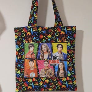 May include: Black tote bag featuring the cast of The Big Bang Theory. The bag has a colourful pattern of science-related icons and the show's logo. The handles are black with the same pattern.