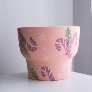 May include: A peach-colored ceramic planter with a festive design. The planter features hand-painted candy canes with white stripes, green sprigs, and red berries, along with white snowflakes. The pot has a wide opening and a stepped base.