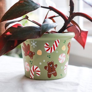 May include: A green ceramic plant pot decorated with gingerbread bears, candy canes, and snowflakes. The pot holds a plant with dark red and green leaves. The pot is sitting on a white surface.