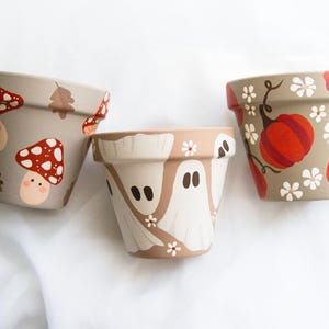 May include: Three hand-painted ceramic flower pots. One pot features red and white mushrooms and a smiling face. Another has white ghosts and flowers. The third pot has orange pumpkins and white flowers. All pots are on a white background.