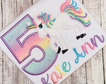 Può includere: Una maglietta bianca con il ricamo di un unicorno con criniera e coda arcobaleno, un corno dorato e zoccoli neri. È presente un grande numero 5 con sfumatura arcobaleno e contorno viola, insieme al nome Kate Ann in scrittura corsiva.