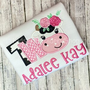 May include: White onesie with a pink and black cow applique with flowers on its head. The onesie also has a pink and black checkered "TWO" and the name "Adalee Kay" in pink.