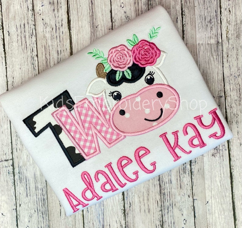 May include: White onesie with a pink and black cow applique with flowers on its head. The onesie also has a pink and black checkered "TWO" and the name "Adalee Kay" in pink.