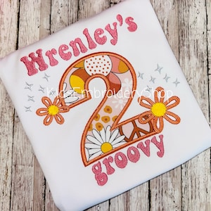May include: White t-shirt with a colorful embroidered design. The design includes the name "Wrenley's" in pink glitter lettering, a large number "2" filled with floral and patterned elements, and the word "groovy" in pink glitter.