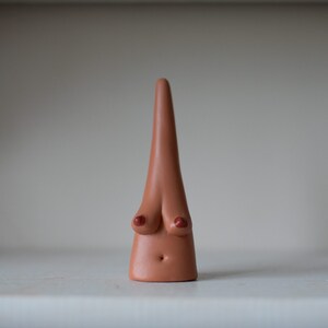May include: A ceramic sculpture of a stylized human form. The sculpture is a light brown color with red accents on the breasts and a small belly button. The form is elongated and conical, with a smooth, matte finish.
