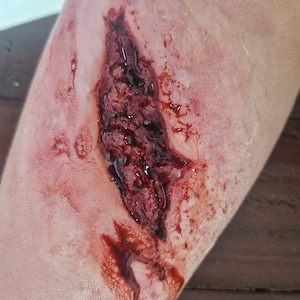 Puede incluir: Una aplicación realista de maquillaje de efectos especiales protésicos que representa una laceración profunda y dentada en la piel. La prótesis presenta tonos de piel texturizados y crudos con sangre simulada roja oscura y brillante.