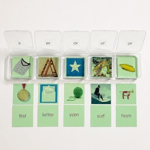 May include: Five clear plastic containers holding cards with the letter combinations ir, er, ar, ur, and or. Below are ten green cards featuring images of a shirt, ladder, star, turtle, corn, medal, letter, yarn, surfer, and horn, with the words first, letter, yarn, surf, and horn.