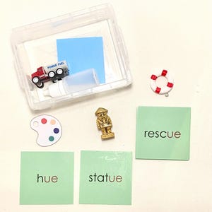 May include: A clear plastic container holds a red and white toy truck, a white bottle, and a blue square. Other items include a miniature gold statue, a palette with colorful paint spots, a white and red life preserver, and green cards with the words "hue", "statue", and "rescue".
