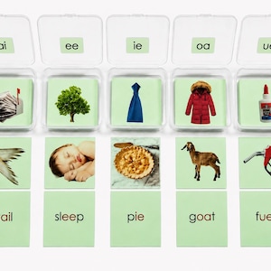 May include: Five clear plastic containers with light green cards featuring vowel teams ai, ee, ie, oa, and ue. Each container holds cards with images and words like tail, sleep, pie, goat, and fuel. The set includes illustrations of a mailbox, tree, tie, coat, glue, bird tail, sleeping baby, pie, goat, and fuel nozzle.