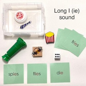 May include: Educational image with items for teaching the long "I" sound. Includes a green toy telescope, a box with a strawberry-patterned lid and a tie, a dice, a fries-shaped pin, and cards with words like "fries", "spies", "flies", and "die".