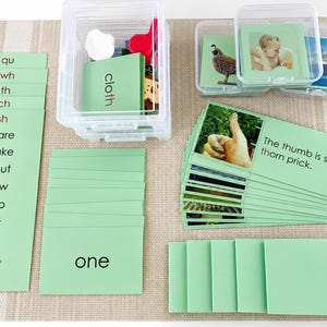 May include: A set of educational phonics cards with light green backgrounds, featuring word lists, individual words like cloth and one, and sentence strips with images. The cards are organised in clear plastic storage containers on a textured mat.