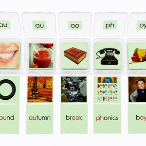 May include: Five clear plastic containers holding educational cards with phonics letter combinations ou, au, oo, ph, and oy. Each set includes a picture card and a word card featuring round, autumn, brook, phonics, and boy with red letter highlights.
