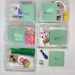 Pode incluir: Seis recipientes de plástico transparente cheios de pequenos objetos e cartões. Cada cartão tem uma palavra: "tail", "three", "oat", "glue", "tie". Os recipientes contêm brinquedos em miniatura, incluindo um barco de brinquedo, uma roda de madeira e um telescópio verde.