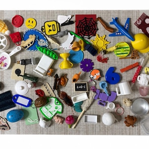 May include: A collection of assorted miniature plastic toys and objects including a blue airplane, a yellow sun, a white frying pan with text, a black spider web, a blue number six, a green pea pod, a small chalkboard with 7-3=?, and various figurines.