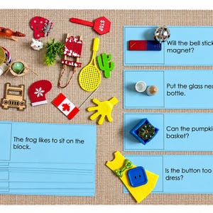 May include: An educational activity set featuring various small plastic and wooden objects including a frog, a medal, a Canadian flag, a stop sign, and a tennis racket alongside blue cards with text prompts like Will the bell stick to the magnet?