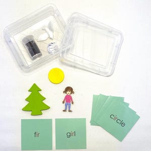 May include: Educational toys and materials, including a green fir tree, a yellow circle, a girl figure, and cards with words like "fir", "girl", and "circle". A clear plastic container holds small items.