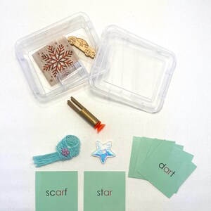 May include: A clear plastic container with a wooden car and a snowflake design. Other items include a gold and orange tool, a blue scarf, a star, and cards with the words "scarf", "star", and "dart".