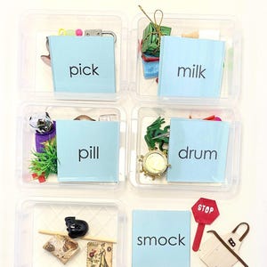 May include: Six clear plastic containers each hold various small objects and a blue card with a word printed in black. The words are "pick", "milk", "pill", "drum", "smock". A red stop sign and a small apron are also present.
