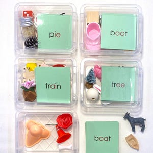 May include: Six clear plastic containers each hold various small objects and a green card with a word printed on it. The words are "pie", "boot", "train", "tree", "boat", and "boat". The objects include a miniature boot, a toy train, and a small goat.