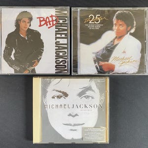 以下が含まれることがあります： プラスチックケースに入ったマイケル・ジャクソンの音楽CD3枚。左上は「Bad」、右上は「Thriller 25」、下は「Invincible」。各ジャケットにはマイケル・ジャクソンのポートレートとアルバムタイトル、テキストが印刷されています。