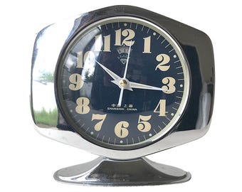 May include: A silver-toned alarm clock with a black face and white numerals. The clock has a diamond-shaped logo and the text "Shanghai, China" on the face.