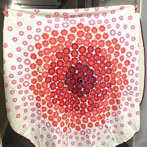 Op de afbeelding: Een ronde witte stoffen kerstboomkleed met een gradiëntpatroon van kleine bloemmotieven. De bloemen verlopen van lichtroze aan de buitenranden naar feloranje en dieprood richting het midden, dat een kleine ronde opening heeft.