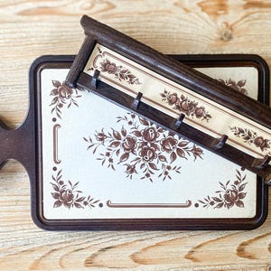 Peut inclure: Planche à découper en bois brun foncé avec une poignée et un porte-ustensiles mural assorti. Les deux articles présentent un motif floral crème. La planche est rectangulaire avec une poignée arrondie. Le porte-ustensiles a des crochets.
