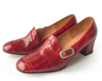 Könnte beinhalten: Ein Paar rote, mit Krokodilprägung versehene Lederschuhe mit Absatz und silberner Schnalle. Die Schuhe haben eine spitze Zehenpartie und einen niedrigen Blockabsatz.