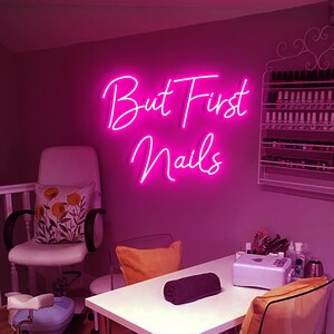 Puede incluir: Un letrero de ne&oacute;n rosa con el texto "But First Nails" colgado en una pared rosa en un sal&oacute;n de manicura. Hay una silla blanca con una almohada floral, una mesa blanca con una toalla negra y una silla transparente con una almohada amarilla.