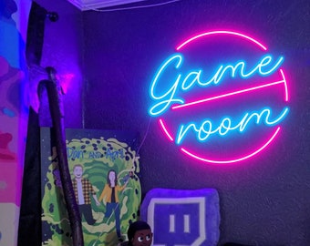 May include: A neon sign with a pink circle and blue text that reads "Game Room". The sign is mounted on a wall with a purple and blue light. There is a purple and white Twitch logo pillow in the foreground.