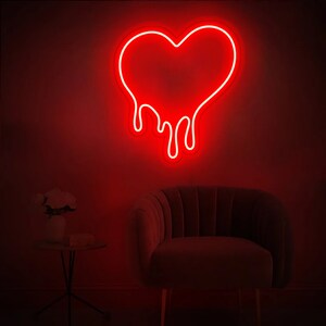 May include: A red neon sign in the shape of a dripping heart. The sign is mounted on a wall in a room with a dark red background. A dark gray armchair is in the foreground.