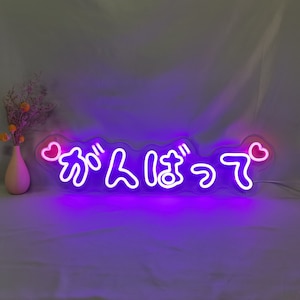 K&ouml;nnte beinhalten: Lilafarbenes Neon-Schild mit dem japanischen Text "ガンバッテ" (ganbatte), was "Gib dein Bestes" oder "Versuch es weiter" bedeutet. Das Schild hat an beiden Enden zwei rosa Herzen.