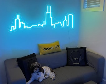 May include: A gray couch with three pillows, one yellow with the text "GAME ON" and two black with a headphone graphic. A small dog is lying on the couch. A blue neon light shaped like a city skyline is on the wall behind the couch.