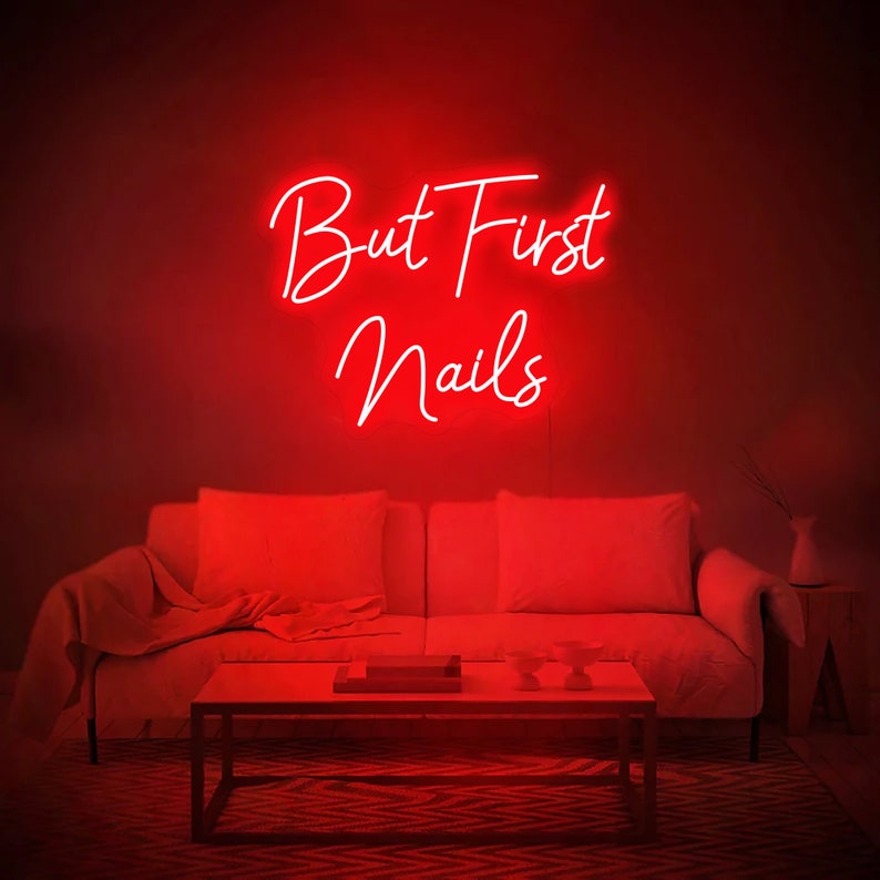 Puede incluir: Un letrero de ne&oacute;n rojo con el texto "But First Nails" colgado en una pared roja sobre un sof&aacute; blanco en una sala de estar.