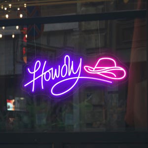 以下が含まれることがあります： 「Howdy」という文字が紫で、ピンクの カウボーイハットが描かれたネオンサイン。窓に吊り下げられています。
