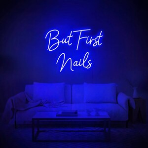 Puede incluir: Un letrero de ne&oacute;n azul con el texto "But First Nails" colgado en una pared sobre un sof&aacute; blanco en una sala de estar.
