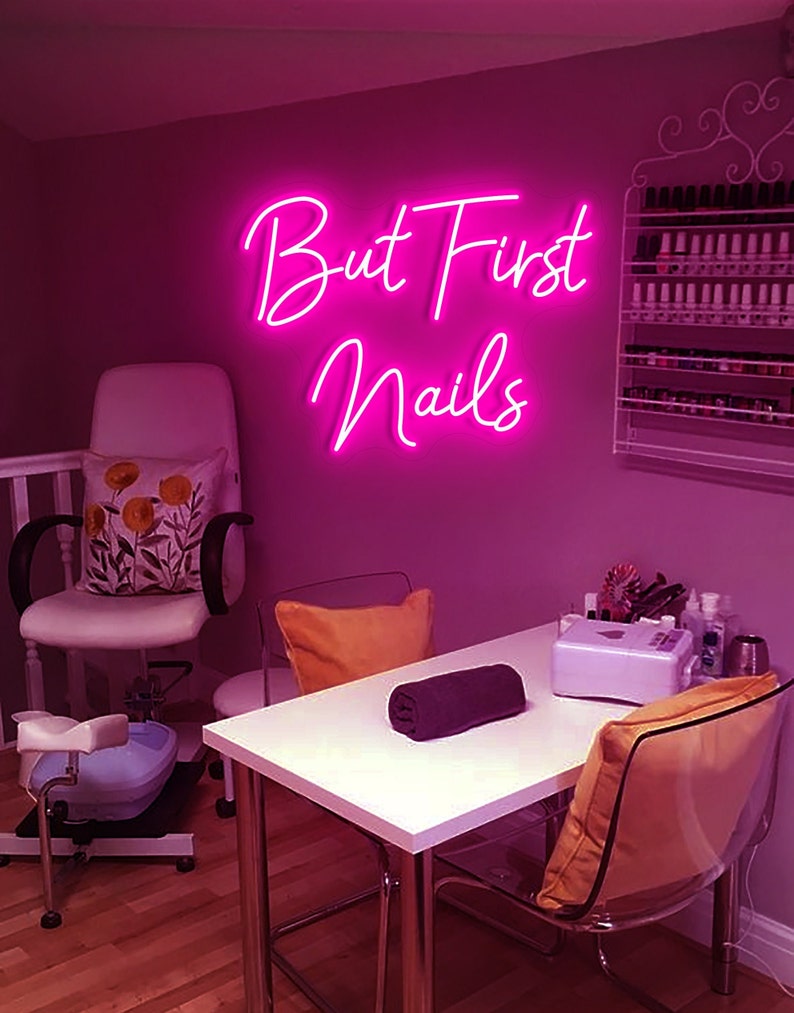 Puede incluir: Un letrero de ne&oacute;n rosa con el texto "But First Nails" colgado en una pared rosa en un sal&oacute;n de manicura. Hay una silla blanca con una almohada floral, una mesa blanca con una toalla negra y una silla transparente con una almohada amarilla.