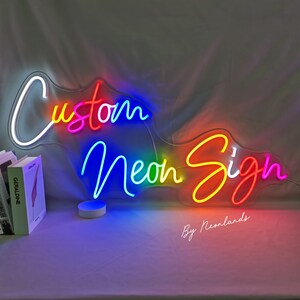May include: A custom neon sign with the words "Custom Neon Sign" in rainbow colours. The sign is lit up and has a white background. The text "By Neonlands" is visible at the bottom of the sign.