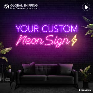 Op de afbeelding: Een aangepast neonbord met de tekst "YOUR CUSTOM Neon Sign" in roze en paars met een gele bliksem. Het bord hangt aan een bakstenen muur voor een zwarte bank.