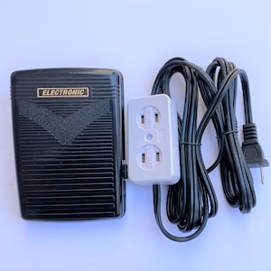 Könnte beinhalten: Schwarzes elektronisches Fußpedal mit einem weißen Netzteil mit zwei Steckdosen, die mit "Motor" und "Licht" beschriftet sind. Das Fußpedal trägt die Aufschrift "Electronic". Das Netzteil ist mit einem schwarzen Stromkabel mit Stecker verbunden.