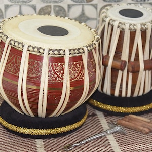 May include: Two Indian tabla drums. The larger drum is red with gold accents and white vertical straps. The smaller drum is brown with white straps. Both drums sit on black cushions with gold trim. A tuning hammer and wooden blocks are in front.