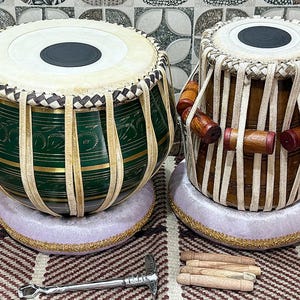 May include: Two tabla drums, traditional Indian percussion instruments. One is dark green with gold accents, the other is brown. Both have white drumheads and are on purple cushions with gold trim. A tuning hammer and tuning pegs are also present.
